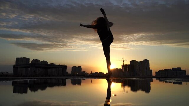 Female flexible dancing in slow motion during sunset on cityscape background with reflection in the water. Concept of freedom and happiness 