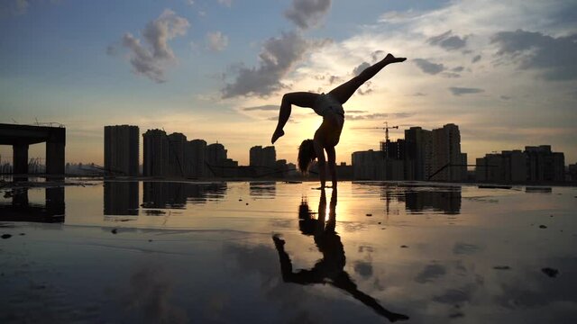 Flexible female gymnast doing handstand and calisthenic with reflection in the water on cityscape background during dramatic sunset in slow motion 