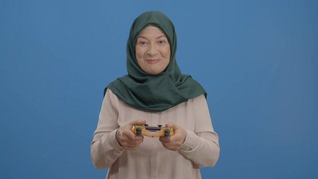 The old woman in a turban playing playstation against a blue background is having fun like crazy. Entertainment concept with Playstation. The concept of having fun together.