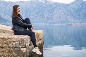 beautiful young brunette woman sits on stone pier and gazes thoughtfully out to sea, beautiful mountain range on background. Single woman travels alone. Restoration of mental health