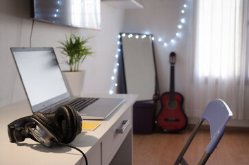 Selective focus of headphones on white desktop and gray laptop, yellow copybook, plant, guitar and mirror defocused.