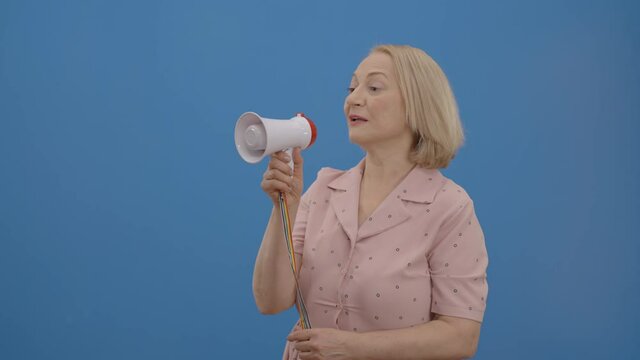 Old Woman Trying To Make Her Voice Heard With A Megaphone In Front Of A Blue Background. The Concept Of Making More Of Your Voice.Slow Motion Video.