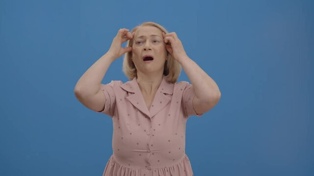 The old woman, who is obviously upset, surprised, panicked, has a headache. Studio shot isolated on blue background.