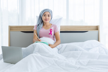Obraz na płótnie Canvas a asian women disease mammary cancer patient with pink ribbon wearing headscarf After treatment to chemotherapy with Take notes in a notebook and laptop on bed In the bedroom at the house,healthcare