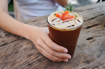 The woman held a glass of strawberry syrup, soda shot, espresso and fresh strawberries in a glass, drank it and put it on a wooden table. 