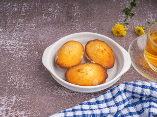 Lemon glaze madeleine on white a dish with a teacup placed on a gray stone background. Madeleine - homemade traditional French small cookie. Space for text. Seashell sweet cakes