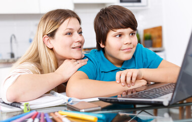 Portrait of smiling mother helping son with homework and having good time in kitchen