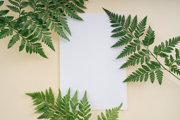 Tropical green leaves with white mock up paper over beige background with copyspace. Top view.