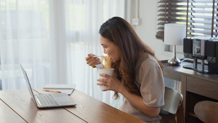 Asian business woman eating instant noodles while working on laptop computer at home office, Happy beautiful young female sitting on desk work overtime doing deadline project, late time business - Powered by Adobe