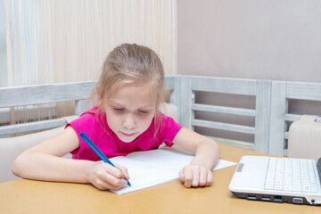 A little girl sits at a table with a laptop and writes homework in a notebook. The child studies at home. Home learning concept