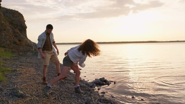 Playful young man and woman in love skipping pebbles across lake water surface on warm spring evening