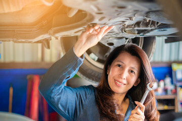Smiling young asian woman mechanic holding wrench standing under a vehicle at the repair garage. Female working underneath a lifted car.