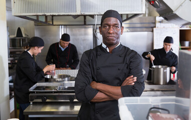 Fototapeta na wymiar Portrait of confident African American chef in restaurant kitchen with busy professional staff