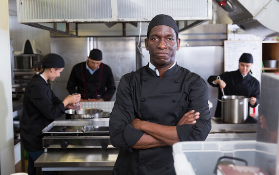 Portrait of confident African American chef in restaurant kitchen with busy professional staff