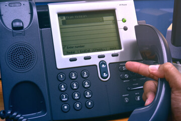 Hand of man holding telephone handset and pressing button on keypad, office desk