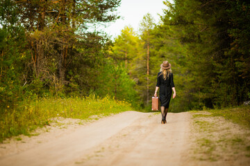 a girl in a black dress and hat walks down the road with a suitcase, selective focus