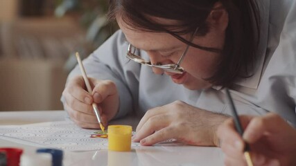 Caucasian man with Down syndrome using paintbrush and gouache while drawing ornament on paper during art lesson with female teacher
