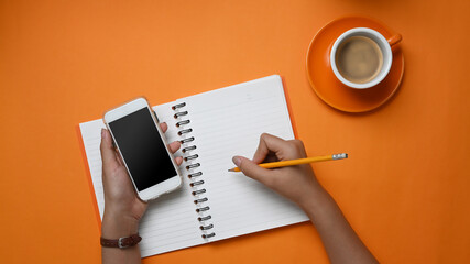 Overhead shot of stylish holding smart phone and making note in notebook on orange background.