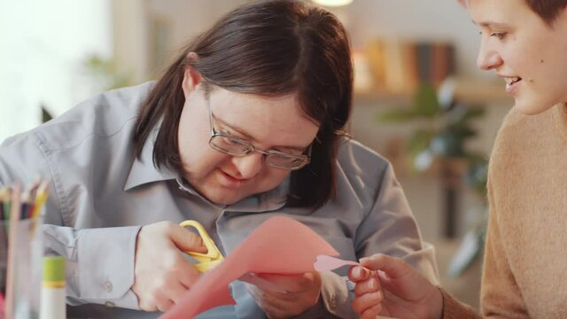 Tilt down shot of Caucasian man with Down syndrome cutting paper with assistance of female teacher during therapeutic art lesson at home