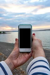 Female hands holding a mobile phone with a picture of sunset on the screen, photographing the sunset on the seascape, back view, close-up, focus on the mobile phone screen, mobile photography.