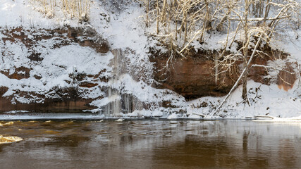 sunny day, landscape with red sandstone cliffs that are snowy with snow, frozen icicles on the cliff wall, icefall on the cliff wall, frozen river, Gauja, Kuku cliffs, Latvia