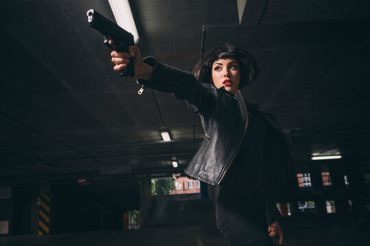 Beautiful brunette sexy spy agent (killer or police) woman in leather jacket and jeans with a gun in her hand running after someone, to catch him on parking