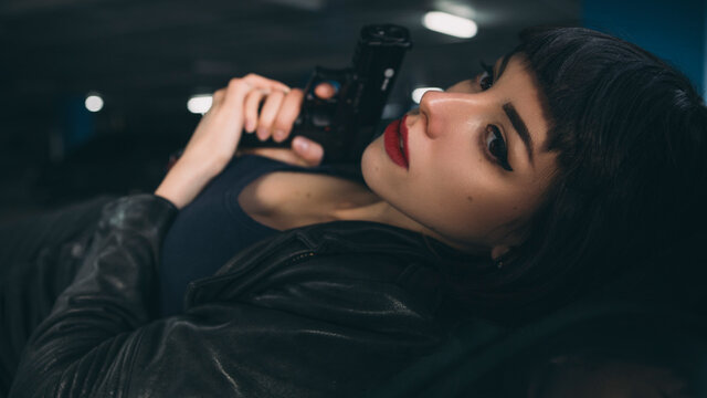 Beautiful brunette sexy spy agent (killer or police) woman in leather jacket and jeans with a gun in her hand running after someone, to catch him on parking