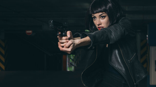 Beautiful brunette sexy spy agent (killer or police) woman in leather jacket and jeans with a gun in her hand running after someone, to catch him on parking
