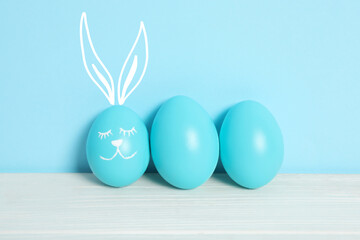 One egg with drawn face and ears as Easter bunny among others on white wooden table against light blue background