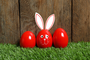 One egg with drawn face and ears as Easter bunny among others on green grass against wooden background