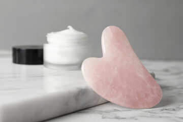 Rose quartz gua sha tool and jar of cream on white marble table, closeup
