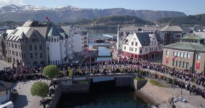 Descending drone footage of marching people amongst the art nouveau buildings and Brosundet bridge of Alesund, Norway's most beautiful town, during national or constitution day, 17th of May.