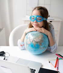 cute blonde girl is sitting at table in front of laptop, holding globe in her hands, has diving goggles or swimming goggles on and dreams about vacation and end of quarantine  and lock down