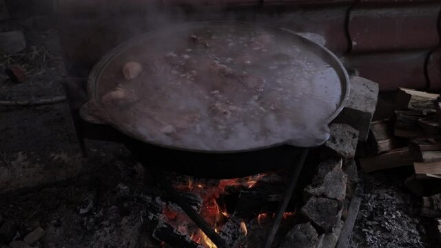 The meat is cooked in a cauldron. Night time. A slow fire burns and steam comes out of the cauldron.