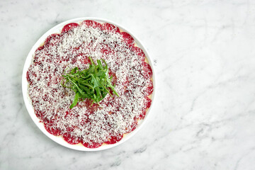 Classic Italian appetizer - veal carpaccio with arugula and parmesan served on a white plate on a marble background. Thinly sliced beef.