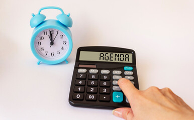 A female hand lies on the calculator with the text on the display AGENDA, on a white background there is an alarm clock