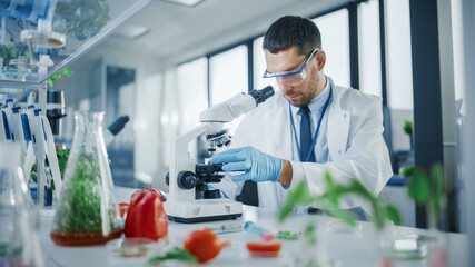 Fototapeta premium Handsome Male Scientist in Safety Glasses Analyzing a Lab-Grown Food Sample Through an Advanced Microscope. Microbiologist Working on Molecule Samples in Modern Laboratory with Technological Equipment