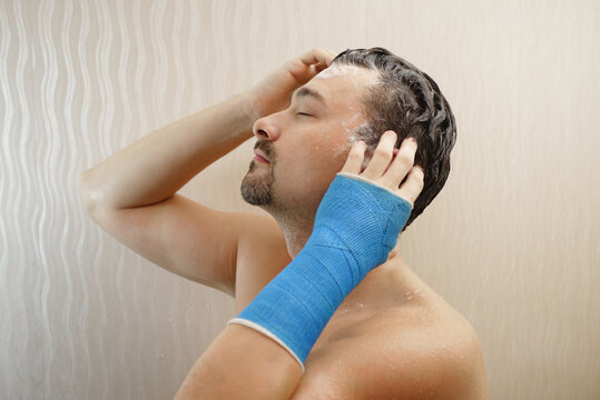 Handsome middle-aged man takes a shower with his hand wrapped in a plaster cast, washes his hair in the bathroom. Broken wrist in modern blue waterproof bandage