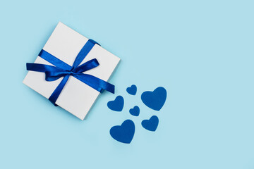 Caja de regalo blanca con un lazo azul junto a corazones azules sobre un fondo celeste liso y...