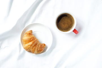 Top view of hot coffee cup beside fresh croissant on white ceramic plate placed on white fabric