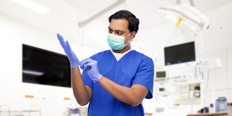 healthcare, profession and medicine concept - indian doctor or male nurse in blue uniform and face protective medical mask putting surgical gloves on over surgery at hospital on background