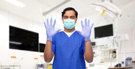 Fototapeta na wymiar healthcare, profession and medicine concept - indian doctor or male nurse in blue uniform, face mask for protection from virus disease and surgical gloves over surgery at hospital on background
