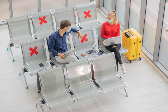 Man and woman travelers wearing protective hygiene mask sitting row seats with social distancing sign in airport lobby and using laptop notebook computer to work while waiting for flight