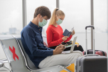 Man and woman travelers wearing protective hygiene masks sitting row seats with social distancing sign in airport lobby. They are checking tickets and documents preparing to check-in and onboard