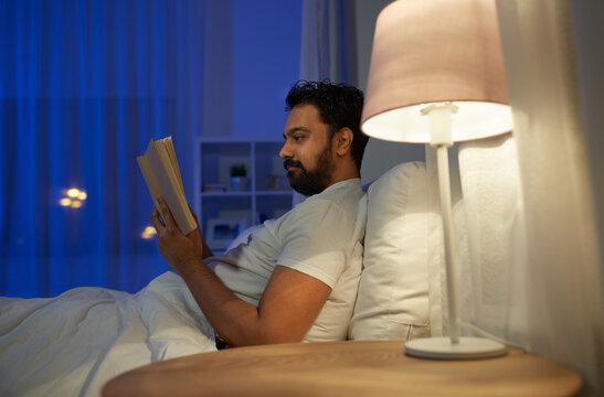 people, bedtime and rest concept - indian man reading book in bed at home at night