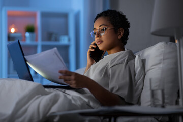 technology, communication and remote job concept - young african american woman with laptop computer and papers calling on smartphone in bed at home at night