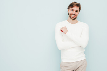 Obraz premium Portrait of handsome smiling hipster lumbersexual businessman model wearing casual white sweater and trousers. Fashion stylish man posing against light blue wall in studio