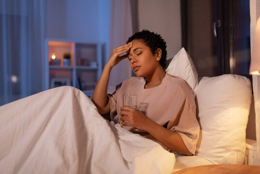 people, bedtime and rest concept - stressed african american woman having headache and drinking water in bed at night