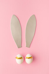 Rabbit ears made of silver foam. Two white eggs with golden lashes. Bunny face concept. Easter pink background.