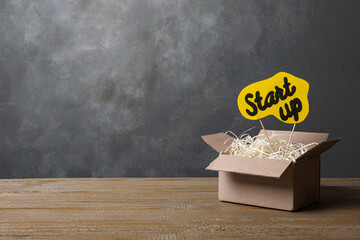 Open cardboard box and sign with words Start Up on wooden table against grey background, space for...